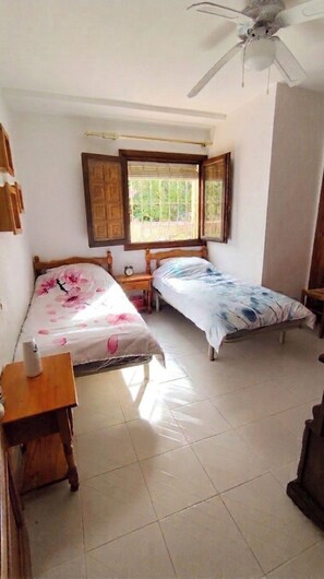 Ein Schlafzimmer mit zwei Einzelbetten, einem Holznachttisch, einem kleinen Tisch und einem Deckenventilator.