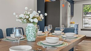 Una tavola imbandita con piatti, ciotole e posate bianche, un vaso con fiori bianchi e una cucina moderna sullo sfondo.