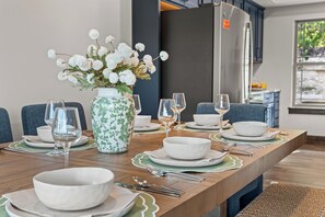 A dining table set with white plates, bowls, and cutlery, a vase with white flowers, and a modern kitchen in the background. - New 6BR Cabin with Hot Tub, Fire Pit, Indoor Pool - Game Table, Shuffleboard (Hollister)