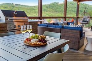 Ein Grill, ein Tisch mit Käseplatte und Weingläsern sowie ein Korbsessel mit blauen Kissen.