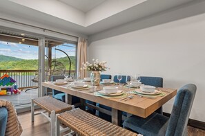 A dining area with a table set for four, chairs, a vase with flowers, and a view of greenery through a large window. - New 6BR Cabin with Hot Tub, Fire Pit, Indoor Pool - Game Table, Shuffleboard (Hollister)