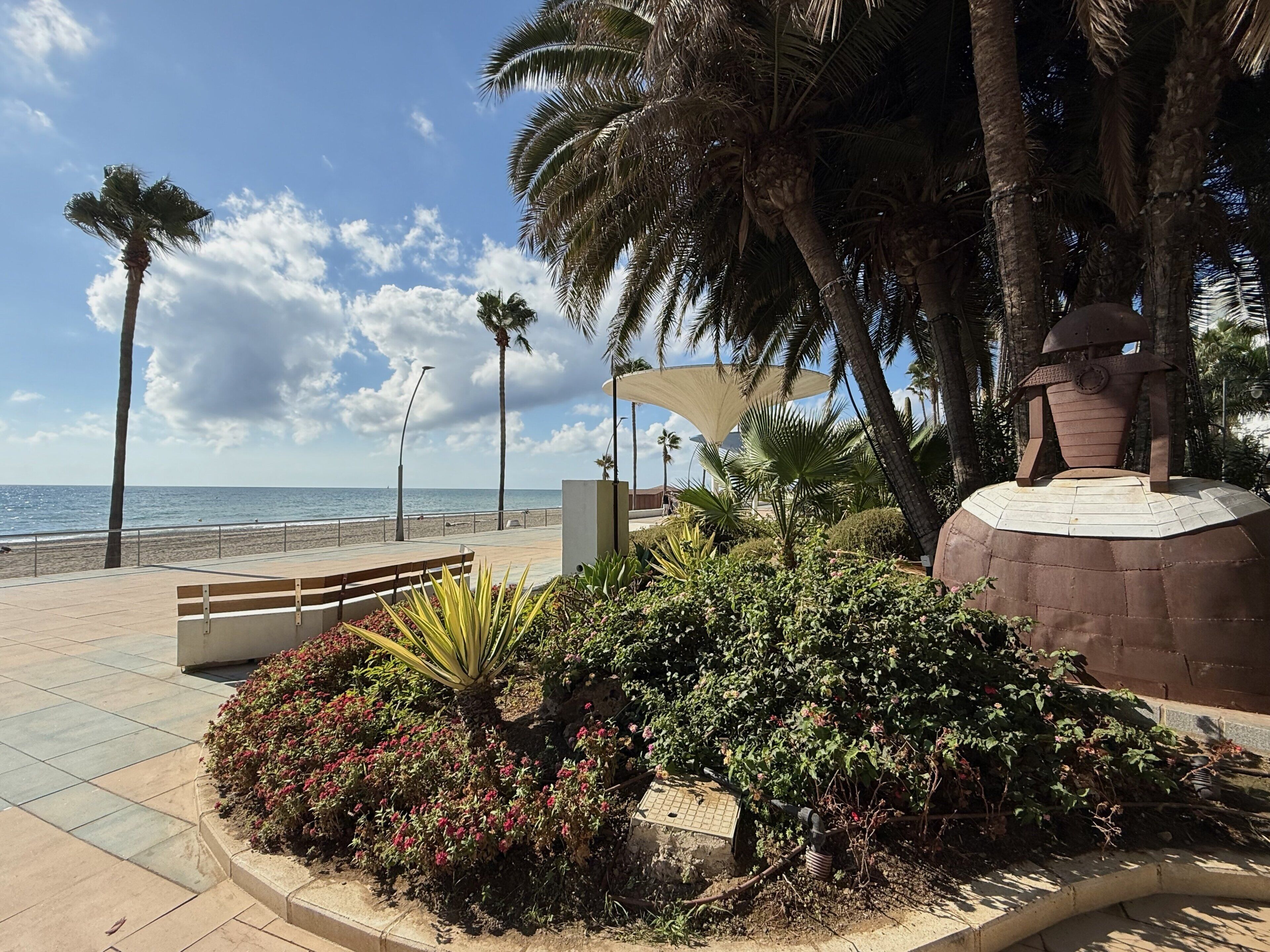 Een boulevard langs het strand met een bankje, palmbomen en een decoratieve constructie.