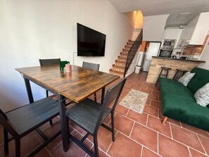 Een eetgedeelte met een houten tafel en stoelen, een groene bank, een aan de muur bevestigde televisie, en een keuken op de achtergrond.