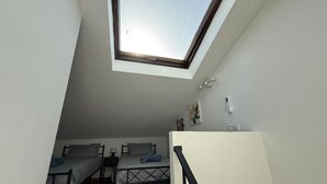 Una camera del dormitorio con due letti, una finestra a lucernario, un muro bianco e un piccolo tavolo con una lampada.