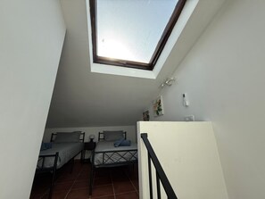 Ein Schlafsaalzimmer mit zwei Betten, einem Oberlicht, einer weißen Wand und einem kleinen Tisch mit einer Lampe.