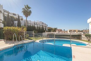A swimming pool with clear blue water, surrounded by a white building, palm trees, and a clear sky. - Calm and bright 1bedroom with Terrace & pool views (Mijas)