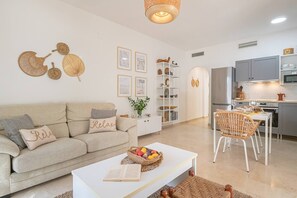 A modern living room with a sofa, a coffee table with a book and fruit basket, a dining area with wicker chairs, and a kitchen with grey cabinets and a TV. - Calm and bright 1bedroom with Terrace & pool views (Mijas)