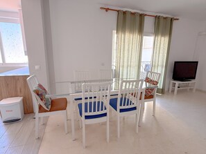 Een eetgedeelte met een glazen tafel, witte stoelen, een tv op een standaard en een raam met gordijnen.