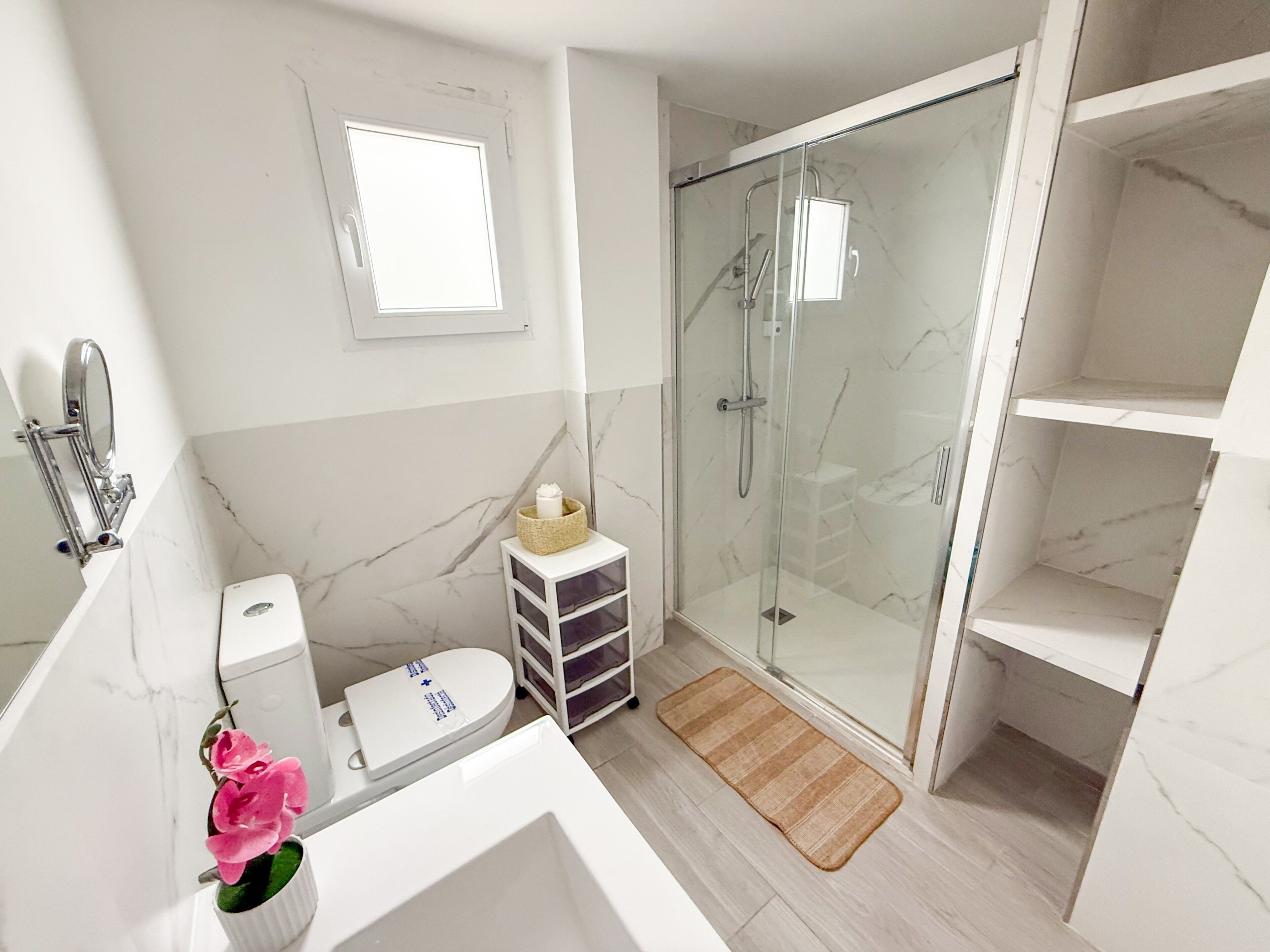 A modern bathroom with a glass-enclosed shower, a white bathtub, a toilet, and a small window.