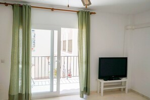 Une pièce dotée d’une porte-fenêtre coulissante, d’un téléviseur posé sur un meuble blanc et d’un balcon avec une rambarde.
