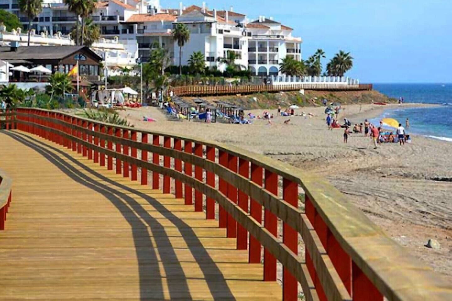 Ein Strand mit einem Holzsteg, Palmen und Gebäuden im Hintergrund.