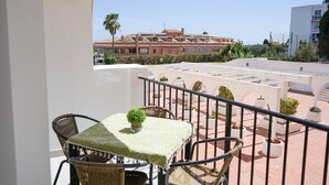 Uma varanda com mesa e cadeiras, um vaso de planta e vista para um prédio com várias janelas e varandas.