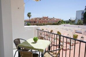 Ein Balkon mit Tisch und Stühlen, einem Blumentopf und Blick auf ein Gebäude mit zahlreichen Fenstern und Balkonen.