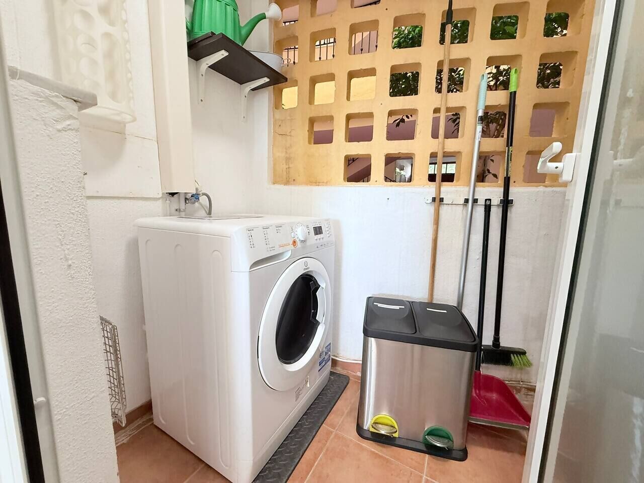 A compact laundry area with a front-loading washing machine, a trash bin, and a broom leaning against the wall.