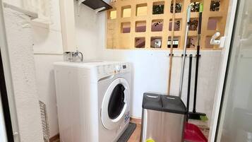 A compact laundry area with a front-loading washing machine, a trash bin, and a broom leaning against the wall.