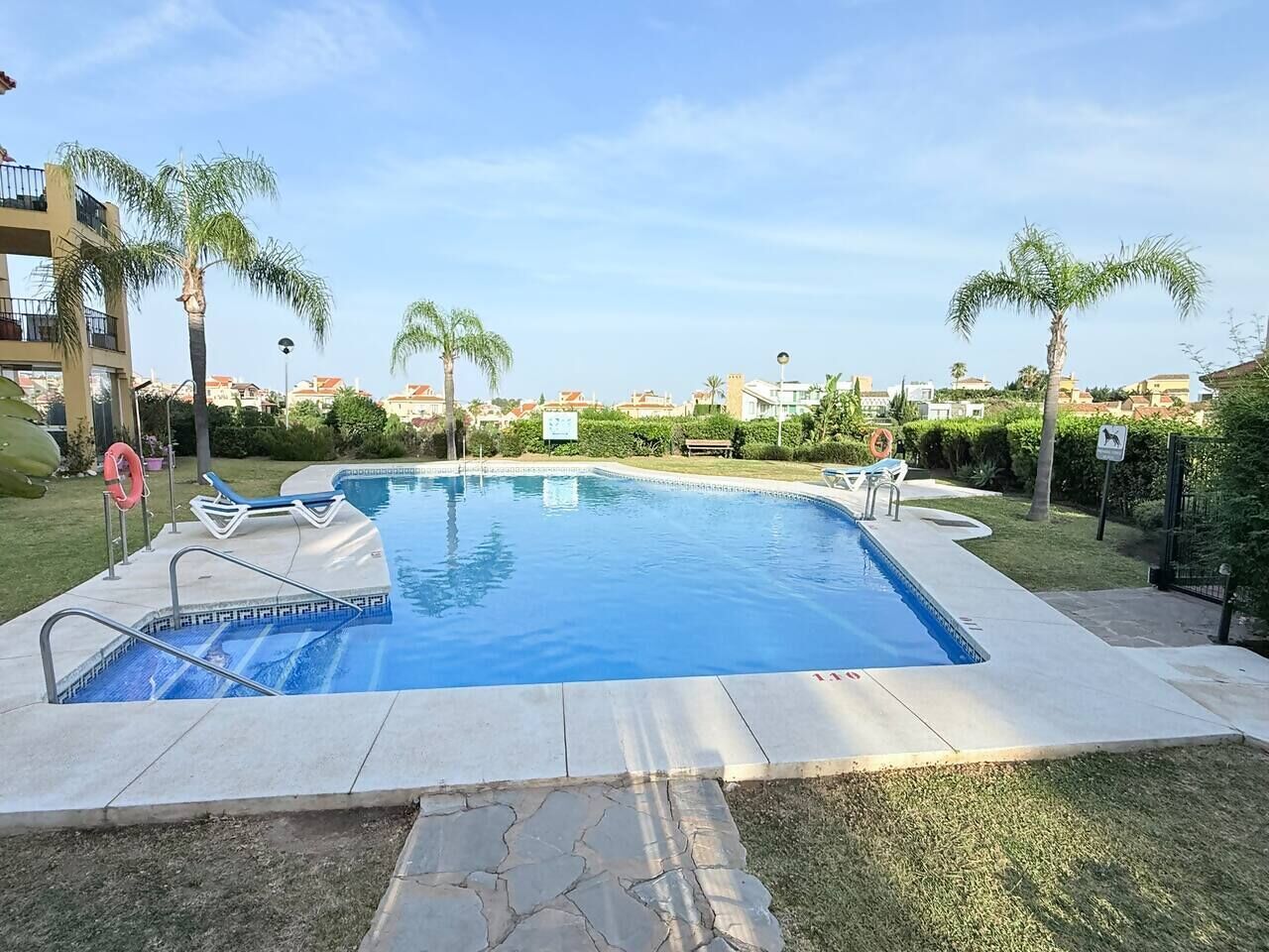 A swimming pool with a tiled deck, surrounded by palm trees and a clear sky.