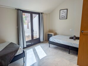 Ein Schlafzimmer mit zwei Betten, einem kleinen Nachttisch, einem gerahmten Bild an der Wand und einer Glastür, die zu einem Außenbereich führt.
