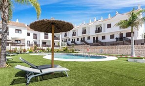 Un espace piscine avec des chaises longues et un parasol en chaume, entouré d’un bâtiment blanc doté de balcons.