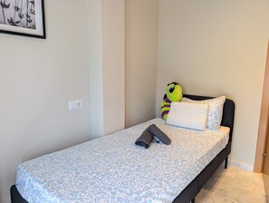 Une petite chambre avec un lit simple, recouverte d’une courtepointe à motifs, avec un oreiller, une serviette enroulée et un jouet en peluche sur le lit.