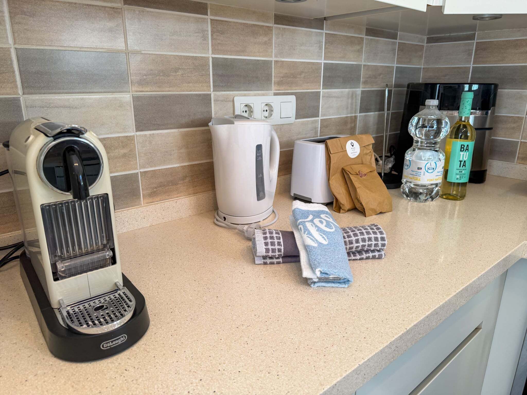 A kitchen counter with a coffee maker, a toaster, a bottle of water, a bottle of olive oil, and a brown paper bag.