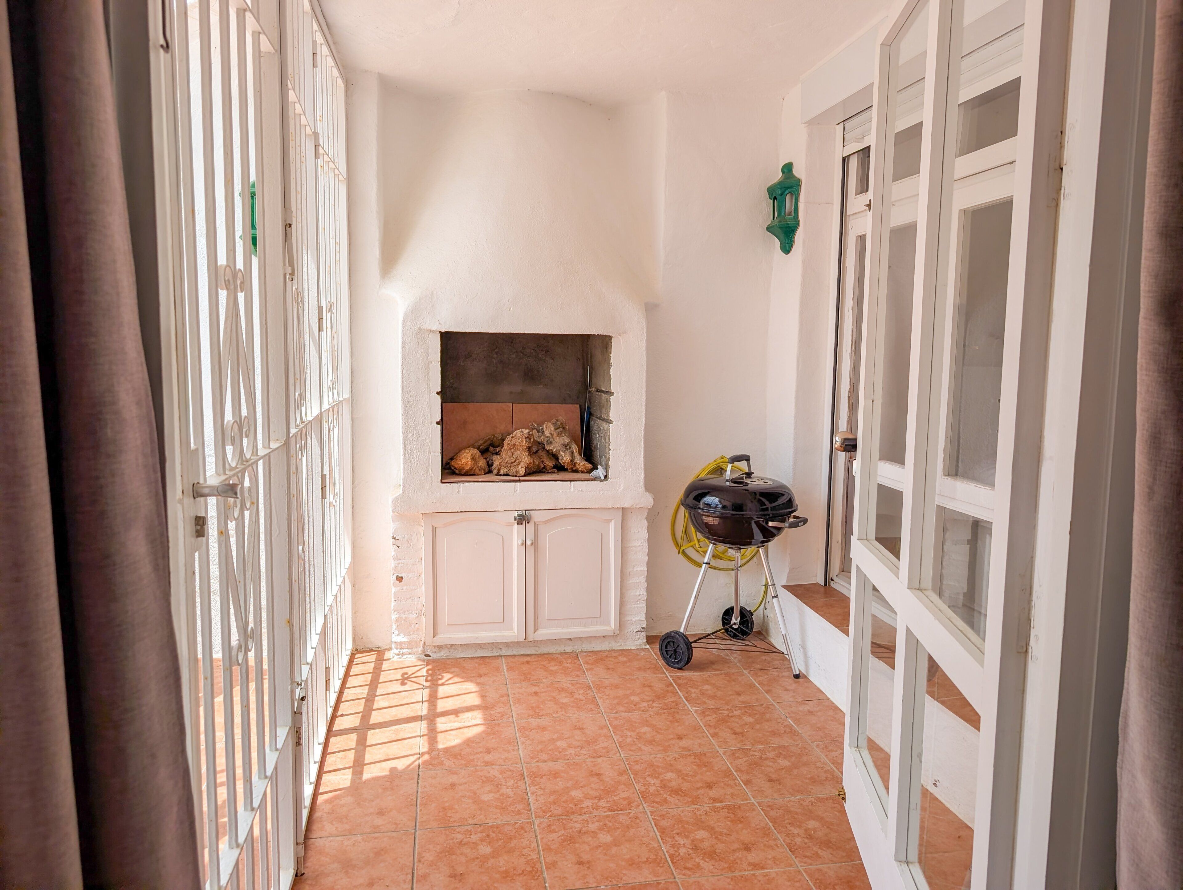 Un couloir avec un sol carrelé, un barbecue équipé d’un tuyau et une lanterne fixée au mur.