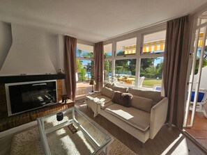 Ein Wohnzimmer mit Kamin, einer Couch und großen Fenstern mit Blick auf Pool und Garten.