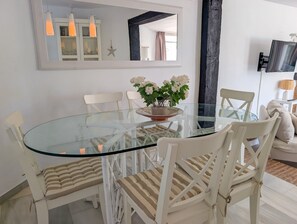 A dining area with a glass table surrounded by white chairs with striped cushions, a decorative plant, and a large mirror above the table. - Calahonda 2BR Garden Apt · Pool · Beach Walk (Mijas)