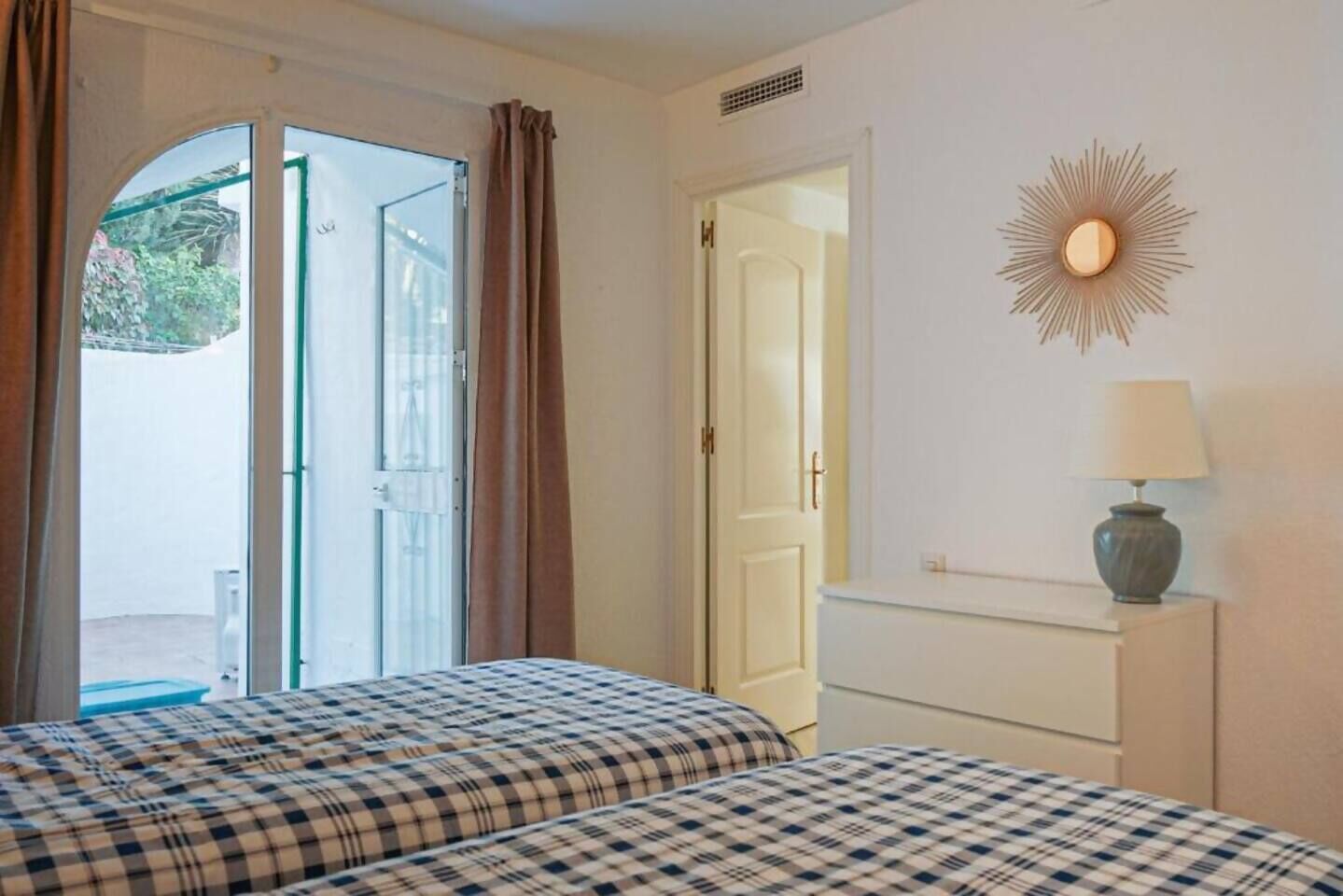 A hotel room with two beds, a window with curtains, a door, and a wall-mounted lamp.