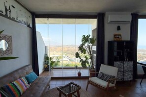 A living room with a sofa, a chair, a small table, and a large window with a view of a cityscape. - Casa Cielo: One bedroom Apt with views in resort (Mijas)