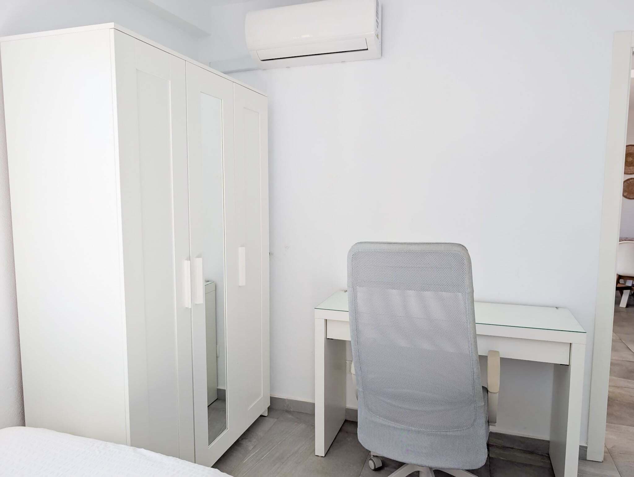 Et værelse med et hvidt skab, et skrivebord med en netlænestol og en aircondition.