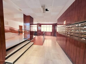 Eine Empfangshalle mit einer Marmortreppe, einer Holzwand mit Briefkästen und Blick durch große Fenster nach draußen.