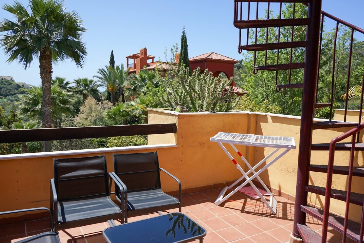 A balcony with two chairs and a table, overlooking a mountainous landscape with palm trees and a red-roofed building.