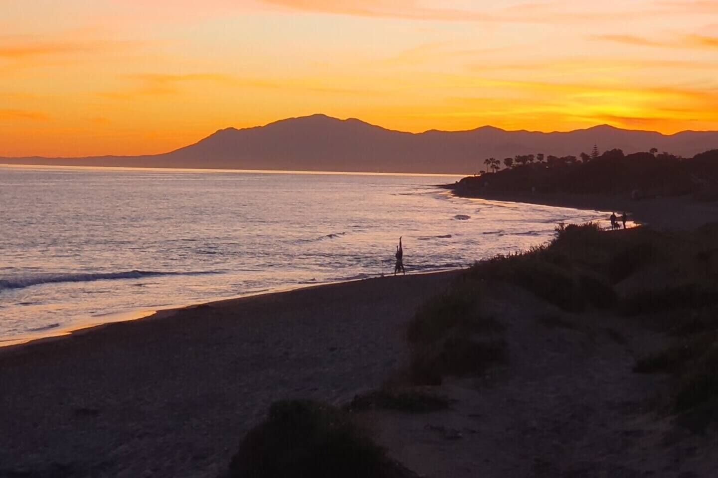 A coastal landscape with a beach, ocean, and distant mountains during sunset.