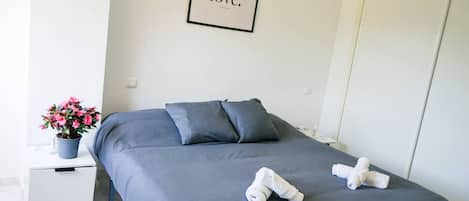 Une chambre à coucher avec un lit, des oreillers, une table de chevet ornée d’un pot de plante et un tableau avec un mot écrit accroché au mur.