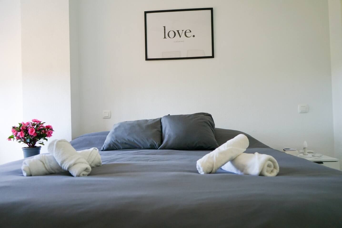 A neatly made bed with pillows and towels, a framed inspirational quote on the wall, and a potted plant on the side.