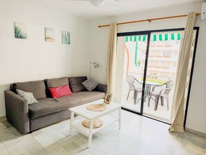 A living room with a sofa, a coffee table, and a sliding glass door leading to an outdoor area with chairs. - Torremolinos: Quiet 1bedroom with pool & sea views (Torremolinos)