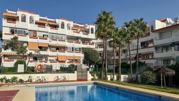 Une piscine à l’eau d’un bleu limpide, entourée d’un espace aménagé avec une clôture, des palmiers et un bâtiment doté de balcons et de fenêtres.