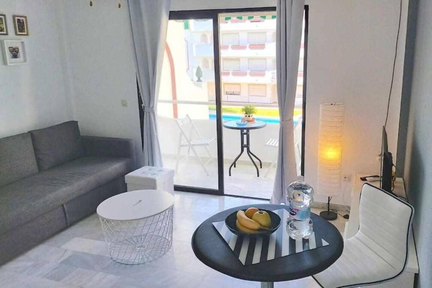 A modern living room with a grey sofa, a round coffee table with fruits and a glass, and a view of a pool and buildings outside.