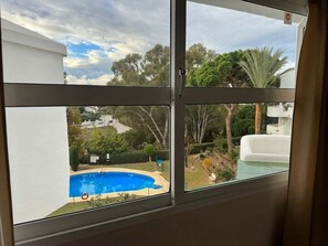 A view of a swimming pool from a window, with a clear sky and surrounding greenery. - Pinos: Serene 2BD w/pool, views & walk to beach (Mijas)