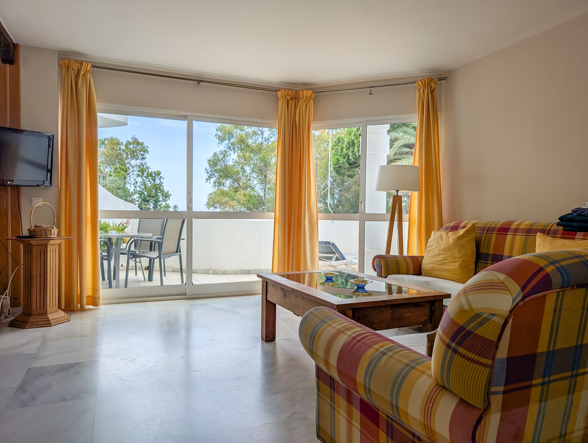 Ein Wohnzimmer mit einer Couch, einem Couchtisch und einem Fernseher. Ein Balkon mit gelben Vorhängen bietet Blick auf Bäume.