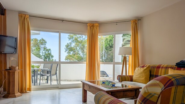 Ein Wohnzimmer mit einer Couch, einem Couchtisch und einem Fernseher. Ein Balkon mit gelben Vorhängen bietet Blick auf Bäume.
