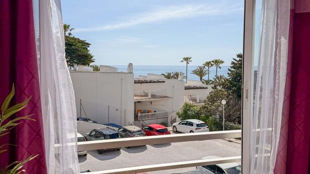 Une vue depuis une fenêtre ornée de rideaux légers, donnant sur un parking où sont garées plusieurs voitures et sur un immeuble à la façade blanche.