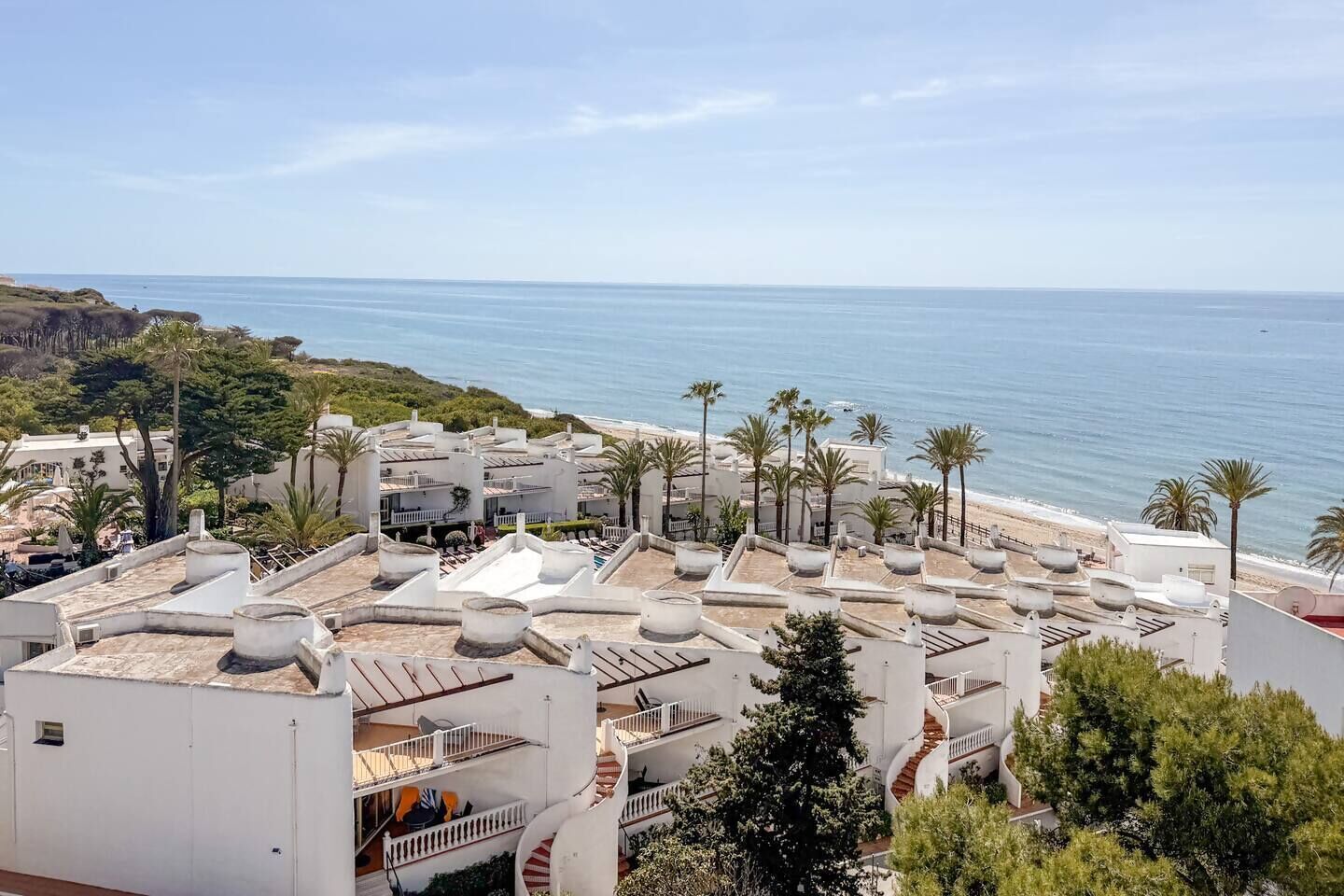 Un hotel costero con varias edificaciones blancas, terrazas en la azotea y palmeras.