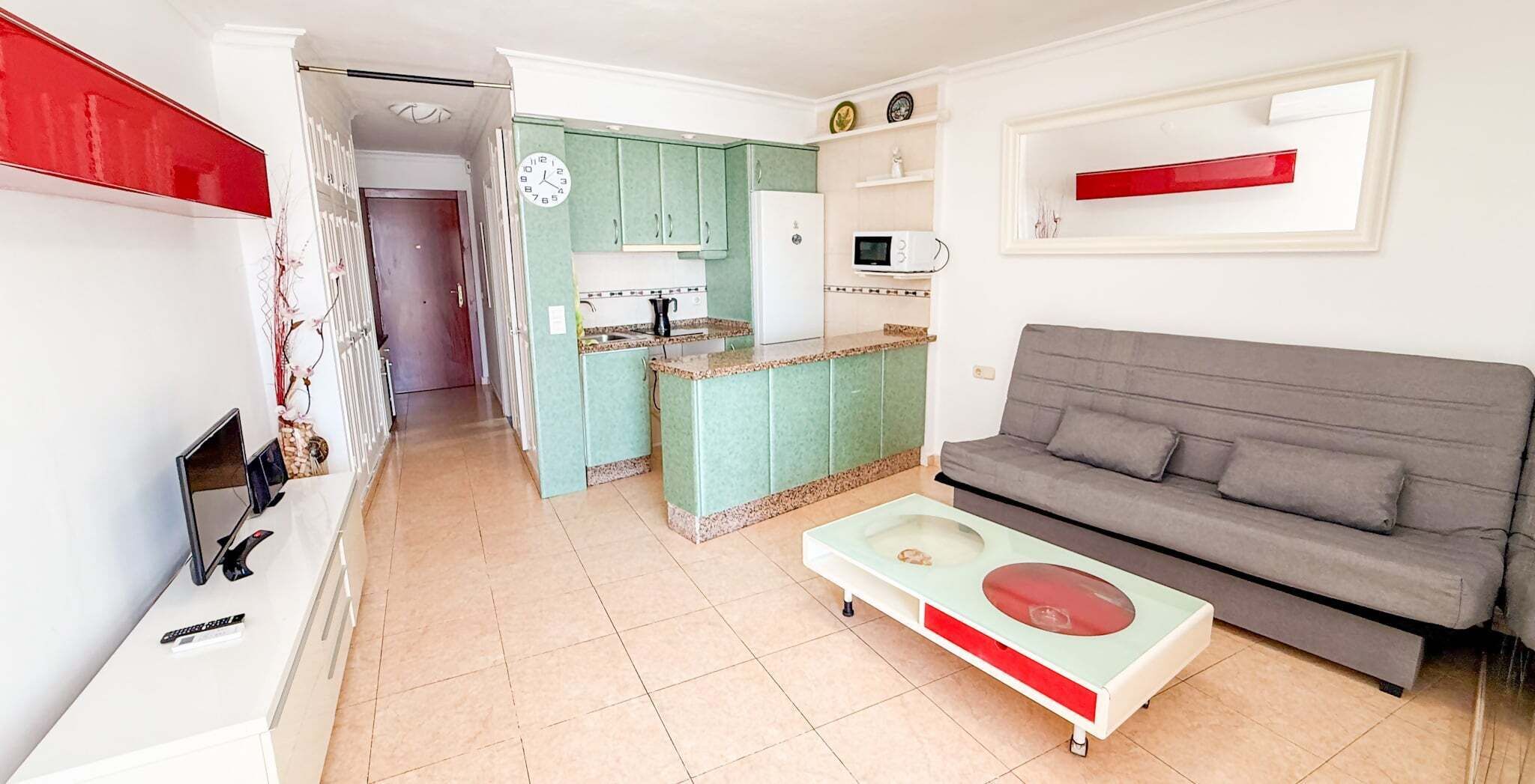 Un espacio habitable compacto con kitchenette, un sofá gris y una pequeña mesa de comedor.