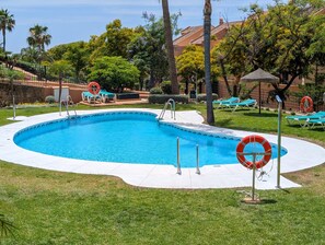 A swimming pool with a curved edge, surrounded by grass and trees, with lounge chairs and a lifebuoy visible. - Casa Lila: Sunny 2bed in Marbella (Marbella)