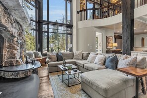 A spacious living area with a stone fireplace, large windows, a high ceiling, and a cozy seating arrangement.