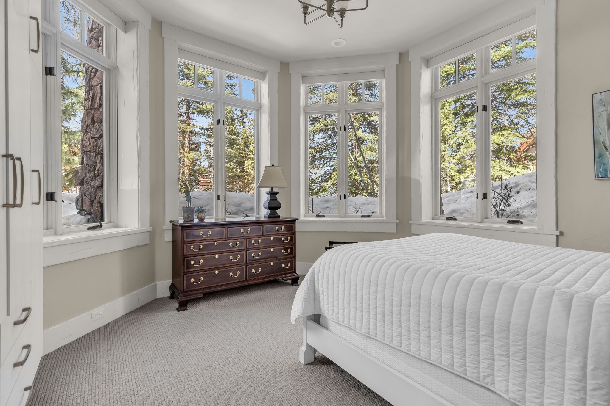 A bedroom with a large bed, a dresser, and multiple windows offering a view of trees and a snowy landscape.