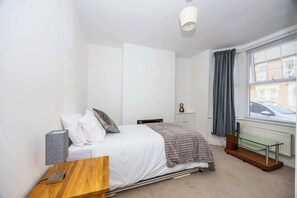 A bedroom with a bed, bedside table, lamp, and a window with curtains. - Spacious 3BR House Near Sixfields, Duston (Northampton)