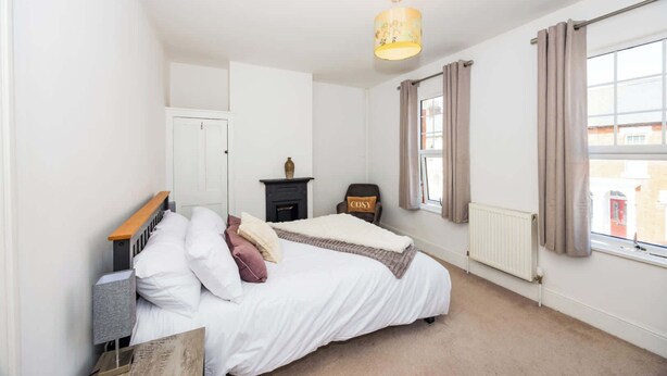 A bedroom with a bed, a nightstand, a lamp, a fireplace, a window with curtains, and a radiator.