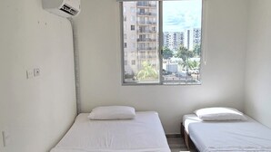 Ein Zimmer mit zwei Betten, einem Fenster mit Blick auf Gebäude, einer Klimaanlage und einem kleinen Tisch.